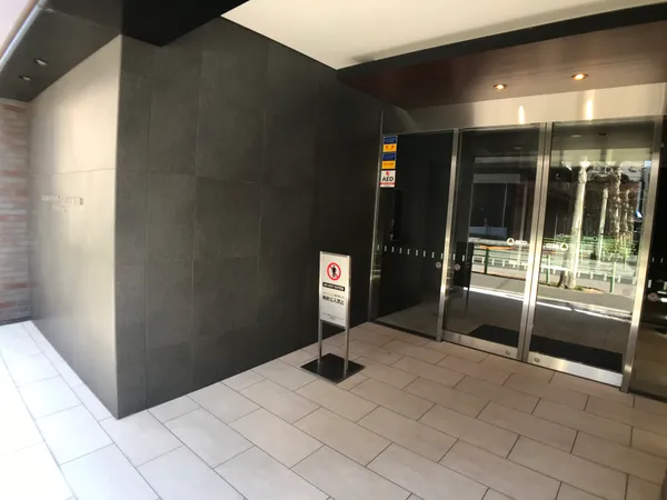 エントランスは、ダークトーンの壁面と白いタイル床が調和した落ち着いた印象です。ガラス扉から自然光が差し込み、明るさと開放感を感じられます。屋根付きのため雨の日でも安心して出入りが可能です。