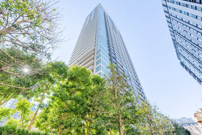 マンションの外観写真です。ガラス張りの外壁とバルコニーが連なるデザインが特徴で、周囲には緑豊かな植栽が広がっています。晴れた空と太陽の光が、建物の高さと開放感を際立たせています。