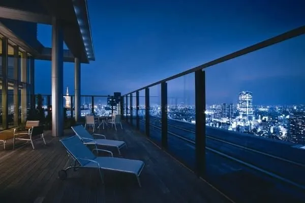 トップラウンジに併設されたスカイテラス。ガラスフェンス越しに都心の夜景が広がり、開放的なウッドデッキにはテーブルやチェアが設置されています。ゆったりとした空間で、都会の景色を眺めることができます。