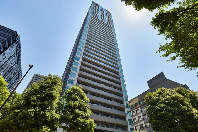 外観写真です。ガラス面が多く、各階にバルコニーが設けられていることが確認できます。周囲には緑豊かな街路樹があり、都市の利便性と自然の調和を感じられる環境です。