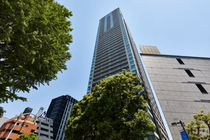 マンションの外観写真です。ガラス張りのバルコニーが連なるデザインが特徴で、周囲には緑豊かな街路樹が広がっています。都市の利便性と自然の調和を感じられる立地です。