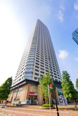 地上高層のタワーマンション外観。ガラス張りの低層部には店舗が入り、周囲は植栽と歩道が整備されています。