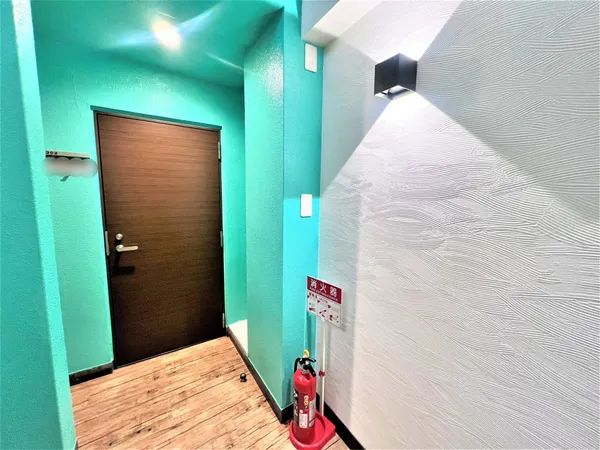 明るいターコイズブルーの壁と木目調の床が印象的な共用廊下です。消火器も設置されており、防災面にも配慮されています。玄関扉はダークブラウンで、落ち着いた雰囲気を演出しています。