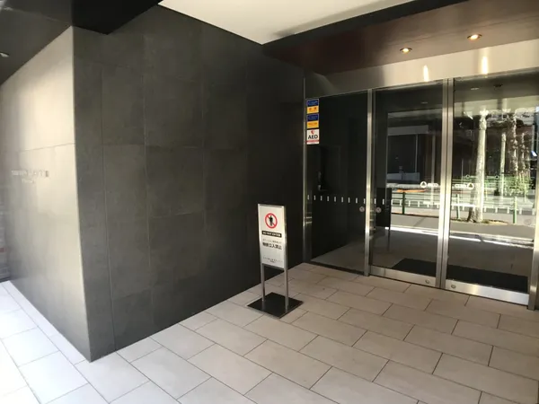 エントランスは、ダークトーンの壁面と白いタイル床が調和した落ち着いた印象です。ガラス扉から自然光が差し込み、明るさと開放感を感じられます。屋根付きのため雨の日でも安心して出入りが可能です。