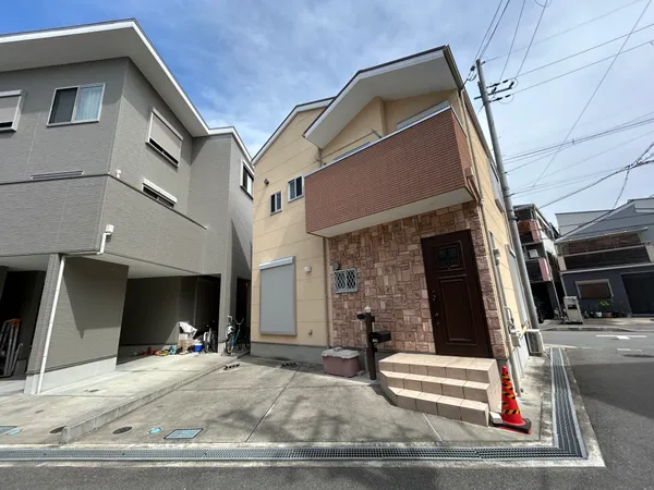 玄関前には階段があり、石調の外壁がアクセントとなっています。前面道路は幅があり、車や自転車の出し入れがしやすい印象です。周囲は住宅が並び、落ち着いた街並みが感じられます。