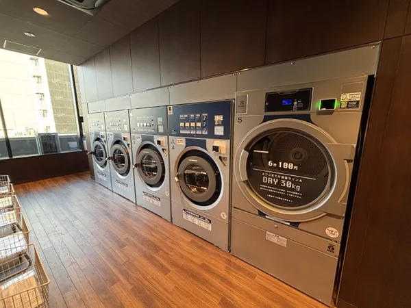共用部のランドリールームの様子です。大型の洗濯機・乾燥機が複数台設置されており、床や壁は落ち着いた色合いの内装でまとめられています。窓からは自然光が入り、明るく清潔感のある空間です。