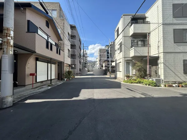 幅員の広い前面道路の様子です。両側に住宅やマンションが並び、日中は明るい空が広がっています。歩道が整えられており、車の通行もスムーズな印象です。周囲の建物との距離感が程よく、落ち着いています。