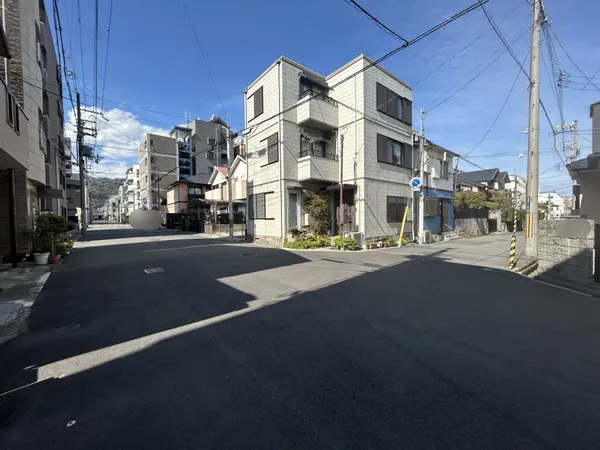 幅員のある交差点に面した住宅街の様子です。周囲は低層～中層の建物が並び、道路は見通しが良く、車や自転車の通行もスムーズな印象で穏やかな生活環境が感じられます。日中は明るく開放的な雰囲気です。