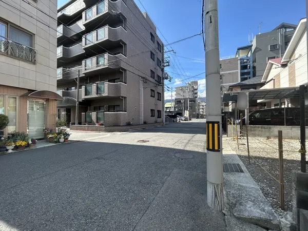 建物前の道路は幅員が広く、車の通行や歩行者の移動もゆとりを感じる印象です。周囲にはマンションや戸建てが並び、落ち着いた住宅街の雰囲気が漂います。